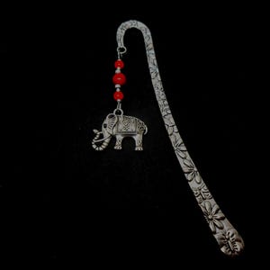 May include: Silver bookmark with a detailed elephant charm and red beads. The bookmark is shaped like a long, thin leaf with a floral design.