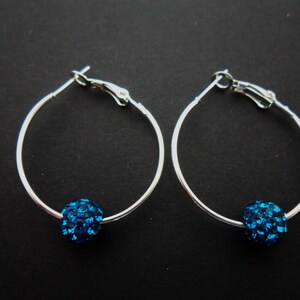 May include: A pair of silver hoop earrings with a bright blue, crystal-covered bead on each. The earrings have a simple, elegant design. The earrings are displayed against a dark background, creating a contrast with the bright blue beads.