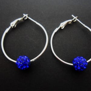 May include: Two silver hoop earrings, each with a bright blue, crystal-encrusted ball. The earrings have a simple, elegant design, ideal for adding a touch of sparkle. The background is a solid black.