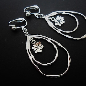 May include: Silver clip-on earrings with a double teardrop design. Each earring has a small, dangling flower charm. The earrings are displayed against a black background, accentuating their metallic shine.