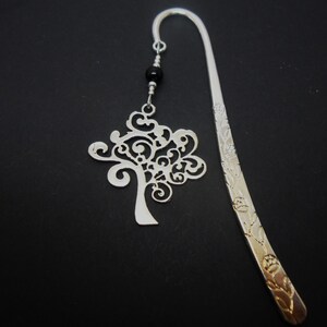 May include: A silver-toned metal hook bookmark featuring a long, curved stem with an engraved floral pattern. A dangling charm shaped like a stylized tree with intricate scrollwork is attached to the top by a small black bead and silver rings.