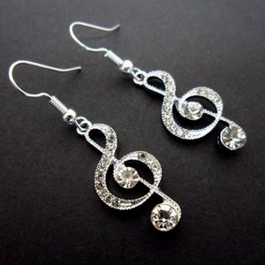 Op de afbeelding: Een paar zilverkleurige bungelende oorbellen in de vorm van een vioolsleutel. Elke oorbel heeft een metalen frame ingelegd met kleine doorzichtige strass-steentjes en is aan de uiteinden versierd met twee grotere, ronde, geslepen stenen.