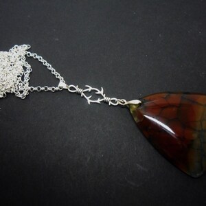 May include: A silver chain necklace featuring a triangular pendant with a polished, multi-colored stone surface displaying deep red, brown, and blue tones with a cracked texture. The pendant connects to the chain via a silver wire-wrapped link.