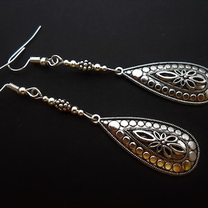 May include: A pair of silver drop earrings. Each earring has a teardrop-shaped pendant with a floral design in the centre, encircled by small circular accents. The earrings have a beaded section and a hook closure.