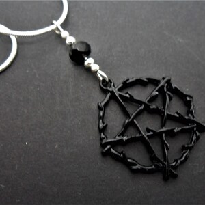 Può includere: Collana con pendente a pentagramma nero. Il pendente è una stella a cinque punte all'interno di un cerchio, con un aspetto strutturato e spinoso. La collana ha una catena argentata e dettagli di perline nere.