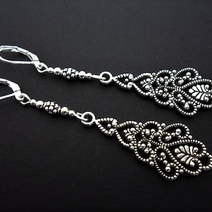 May include: A pair of silver dangle earrings with intricate filigree designs. The earrings are made of small beads and have a teardrop shape.