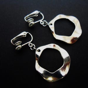May include: A pair of silver-toned clip-on earrings featuring irregular, wavy-edged circular pendants. Each earring consists of a polished metal clip mechanism connected by a small jump ring to a flat, open-center, organic-shaped dangle charm.
