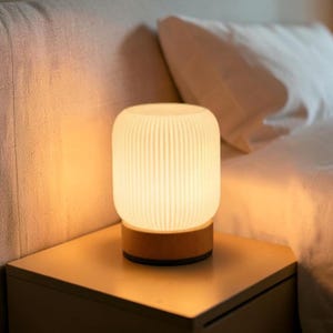 May include: A cylindrical table lamp featuring a translucent, vertically ribbed white shade that emits a warm, soft glow. The lamp is supported by a smooth, circular wooden base with a dark trim at the bottom, providing a stable foundation.