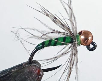 May include: Close-up of a handmade fishing fly. The fly features a copper-colored bead head, a green body with black stripes, and delicate gray and brown feathers. The hook is black and held by a textured, dark gray material.