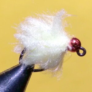 May include: Close-up of a handmade fishing fly. The fly features a fluffy, light-yellow body with a red bead head and a black hook. The fly is designed for attracting fish in freshwater environments.
