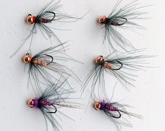 May include: Six handmade fishing flies. Each fly features a copper-colored bead head, a black hook, and a body wrapped with various colors of thread, including red, orange, and purple. The flies have long, gray, feathery extensions.