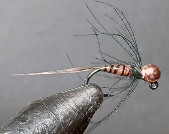 May include: Close-up of a fishing fly with a copper-colored bead head and a black hook. The fly's body is brown with dark brown stripes, and it has black feather-like extensions. The fly is held by a dark, textured tool.
