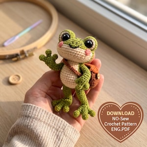 May include: A small green crocheted frog toy with a cream-colored belly, pink cheeks, and large black eyes. The frog wears a brown crocheted backpack with a small wooden button. Text on the image reads: DOWNLOAD NO-Sew Crochet Pattern ENG.PDF.