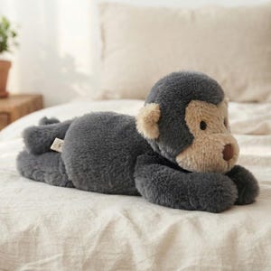 May include: A plush monkey toy featuring dark gray fur on its body and limbs, with a contrasting light beige face, ears, and muzzle. The stuffed animal has a small brown nose and is depicted in a lying position with its front paws extended forward.