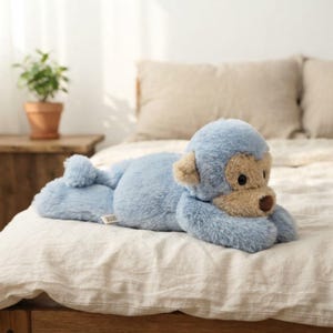 May include: A plush monkey toy with soft light blue fur and a contrasting beige face. The stuffed animal features dark brown eyes and a small brown nose, with a small white fabric tag visible on its side. It is lying in a prone position.