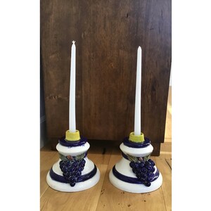 May include: Two white ceramic candle holders with blue and gold accents. Each holder features a grape cluster design and a yellow candle.