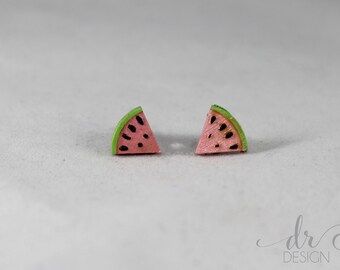 Könnte beinhalten: Zwei Ohrringe in Form von Wassermelonenstücken. Jeder Ohrring ist ein rosa Dreieck mit schwarzen Samendetails und einer grünen Schale. Die Ohrringe sind klein, wahrscheinlich Ohrstecker, und sind vor einem weißen Hintergrund platziert.
