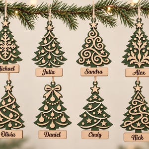 May include: Eight hanging Christmas tree ornaments made of dark green and light wood. Each tree features a unique cutout design, a star topper, and a rectangular wooden name tag at the base with the names Michael, Julia, Sandra, Alex, Olivia, Daniel, Cindy, and Nick.