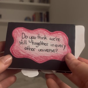 Può includere: Un cartoncino rettangolare nero con una forma rosa irregolare disegnata a mano al centro. All'interno della forma rosa, il testo recita: Do you think we're still together in every other universe? Sono visibili inserti di carta bianca sui bordi.