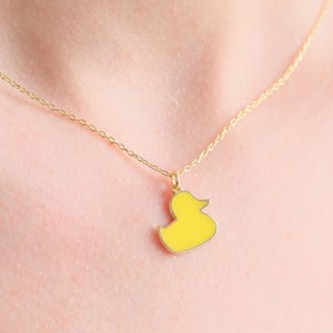 May include: A delicate gold chain necklace featuring a small, bright yellow enamel duck pendant. The pendant is shaped like a classic rubber duck in profile, suspended from a thin, polished gold-tone metal chain that rests against the neck.