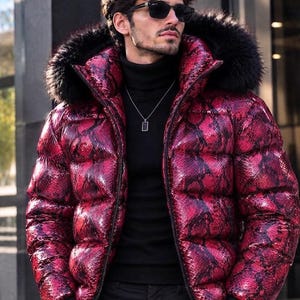May include: A puffer jacket featuring a vibrant red and black snakeskin pattern, a high collar, and a hood trimmed with thick black faux fur. The jacket is worn over a black turtleneck sweater, paired with a silver rectangular pendant necklace.