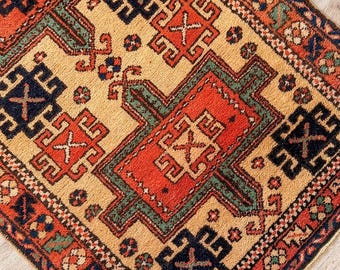 Puede incluir: Una alfombra rectangular con un patrón geométrico complejo en tonos naranja, azul oscuro y verde sobre un fondo color crema. La alfombra muestra motivos repetitivos con bordes intrincados y una construcción de pelo tejido texturizado.