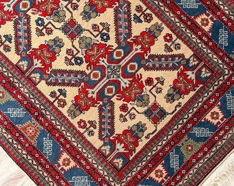 Puede incluir: Una alfombra rectangular con un complejo patrón geométrico y floral en tonos rojo, azul, crema y verde. La alfombra presenta un borde decorativo con motivos repetitivos y flecos blancos cortos a lo largo de los bordes del tejido.