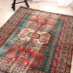 Puede incluir: Una alfombra rectangular con un complejo patr&oacute;n geom&eacute;trico en tonos rojo intenso, verde bosque, azul marino y crema. La alfombra tiene un borde decorativo ancho con motivos intrincados y flecos cortos en los extremos de la tela.
