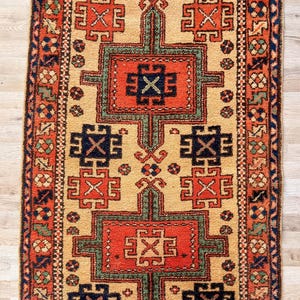 K&ouml;nnte beinhalten: Ein rechteckiger Teppich mit beigem Hintergrund und einem zentralen Design aus zwei gro&szlig;en, rot-gr&uuml;nen geometrischen Medaillons. Der Teppich enth&auml;lt kleinere dunkelblaue und rote geometrische Motive, eingefasst von einer breiten roten Bord&uuml;re.