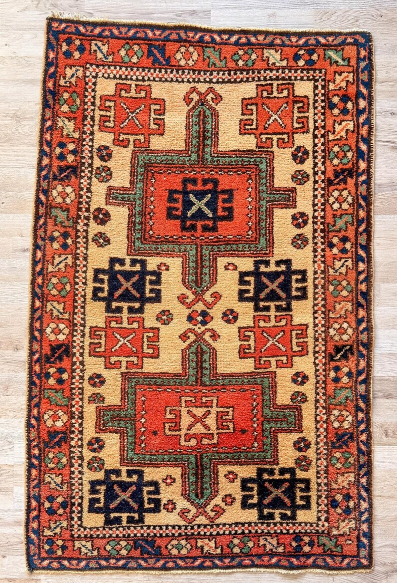 K&ouml;nnte beinhalten: Ein rechteckiger Teppich mit beigem Hintergrund und einem zentralen Design aus zwei gro&szlig;en, rot-gr&uuml;nen geometrischen Medaillons. Der Teppich enth&auml;lt kleinere dunkelblaue und rote geometrische Motive, eingefasst von einer breiten roten Bord&uuml;re.