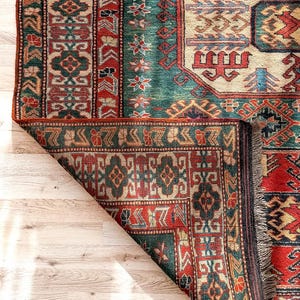 Puede incluir: Una alfombra rectangular con un complejo patr&oacute;n geom&eacute;trico en tonos rojo intenso, verde azulado, crema y azul marino. La alfombra tiene una esquina doblada que revela el reverso, un borde tejido grueso y flecos cortos de color claro.