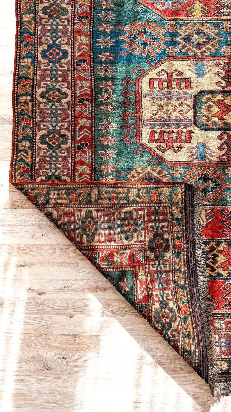 Puede incluir: Una alfombra rectangular con un complejo patr&oacute;n geom&eacute;trico en tonos rojo intenso, verde azulado, crema y azul marino. La alfombra tiene una esquina doblada que revela el reverso, un borde tejido grueso y flecos cortos de color claro.