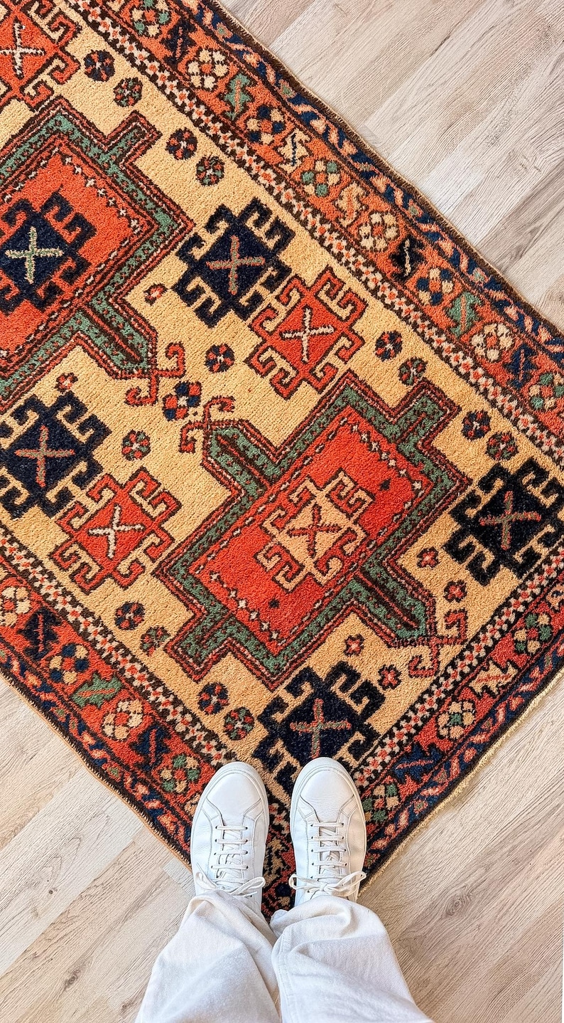 K&ouml;nnte beinhalten: Ein rechteckiger Teppich mit einem komplexen geometrischen Muster in Orange-, Dunkelblau- und Gr&uuml;nt&ouml;nen auf cremefarbenem Grund. Der Teppich zeigt sich wiederholende Motive mit komplizierten R&auml;ndern und einer strukturierten, gewebten Floroberfl&auml;che.