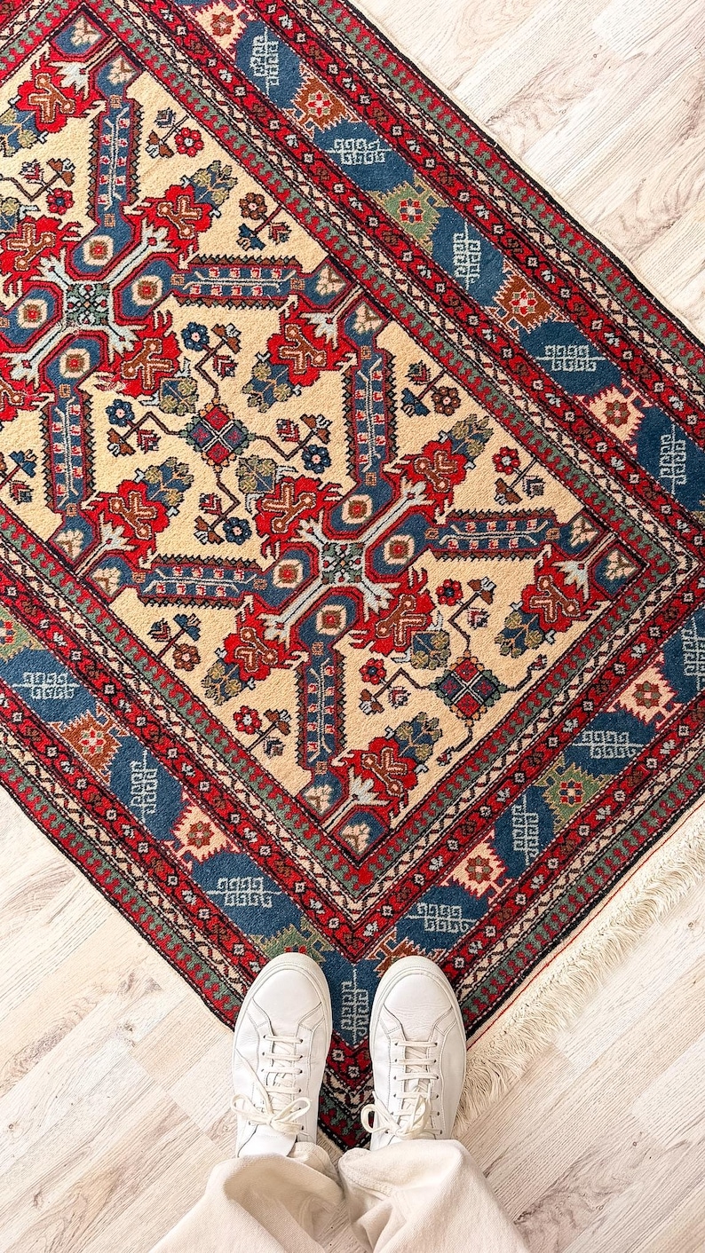 Op de afbeelding: Een rechthoekig vloerkleed met een complex geometrisch en bloemmotief in tinten rood, blauw, cr&egrave;me en groen. Het kleed heeft een decoratieve rand met herhalende motieven en korte witte franjes langs de randen van het kleed.