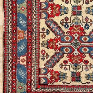 Op de afbeelding: Een rechthoekig vloerkleed met een cr&egrave;mekleurig veld en een geometrisch patroon in rood, blauw en groen. De rand bestaat uit meerdere rode en blauwe banden met complexe florale en geometrische motieven, afgewerkt met een korte franje.