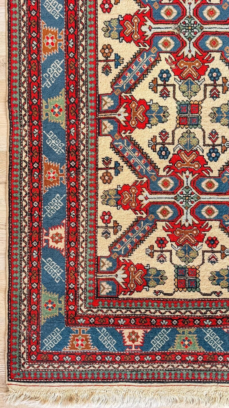 Op de afbeelding: Een rechthoekig vloerkleed met een cr&egrave;mekleurig veld en een geometrisch patroon in rood, blauw en groen. De rand bestaat uit meerdere rode en blauwe banden met complexe florale en geometrische motieven, afgewerkt met een korte franje.