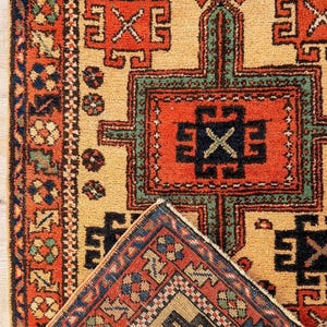 K&ouml;nnte beinhalten: Ein rechteckiger Teppich mit einem geometrischen Muster in den Farben Orange, Beige, Dunkelblau und Gr&uuml;n. Das Design umfasst sich wiederholende kreuzartige Motive und komplexe Bord&uuml;ren. Eine Ecke des Teppichs ist umgeschlagen und zeigt die Unterseite.