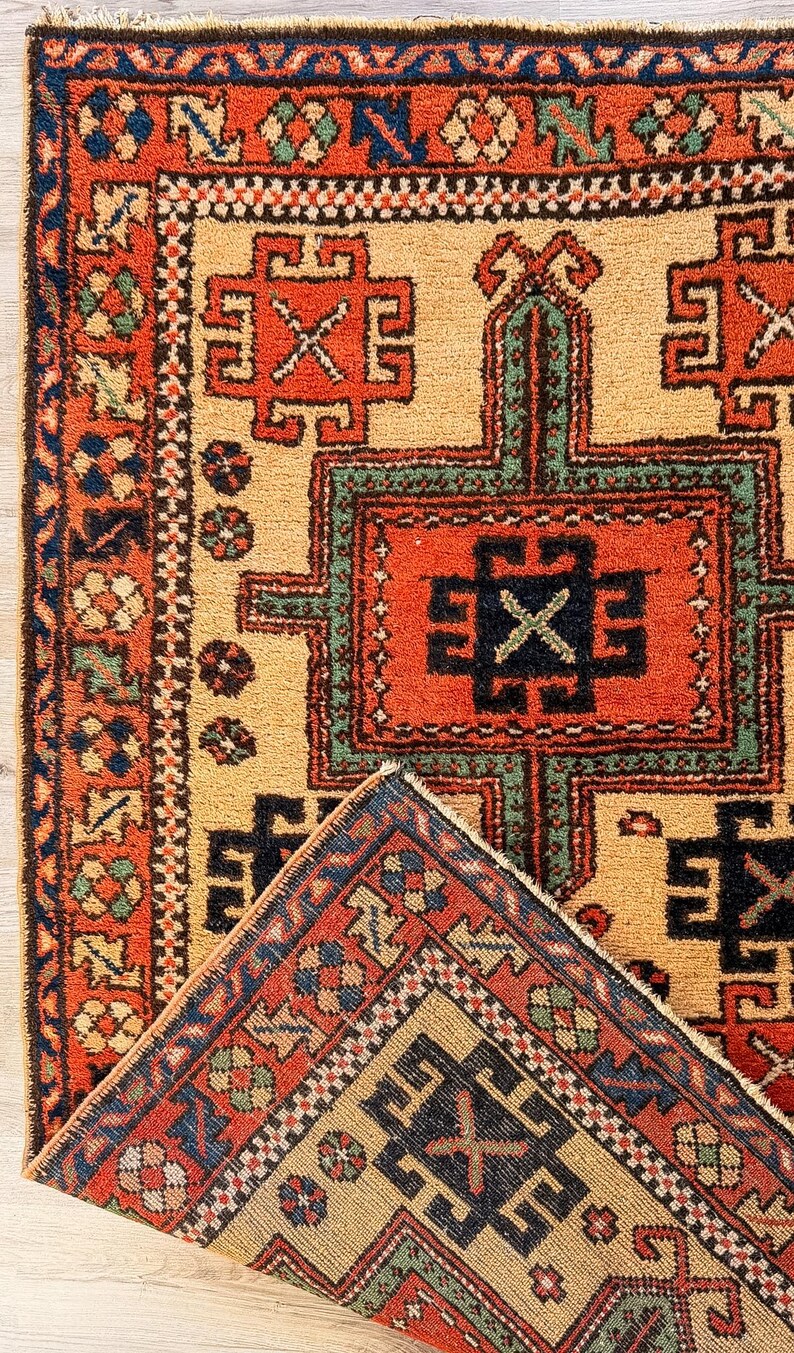 K&ouml;nnte beinhalten: Ein rechteckiger Teppich mit einem geometrischen Muster in den Farben Orange, Beige, Dunkelblau und Gr&uuml;n. Das Design umfasst sich wiederholende kreuzartige Motive und komplexe Bord&uuml;ren. Eine Ecke des Teppichs ist umgeschlagen und zeigt die Unterseite.