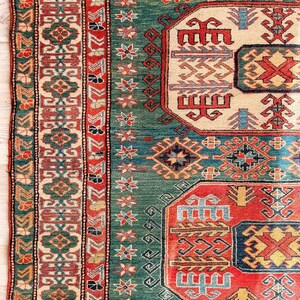 Puede incluir: Una alfombra rectangular con un campo verde intenso y medallones geom&eacute;tricos repetitivos en rojo, crema y azul. El borde muestra intrincados patrones florales y en zigzag en tonos rojos, verdes y crema, rematada con un fleco corto.