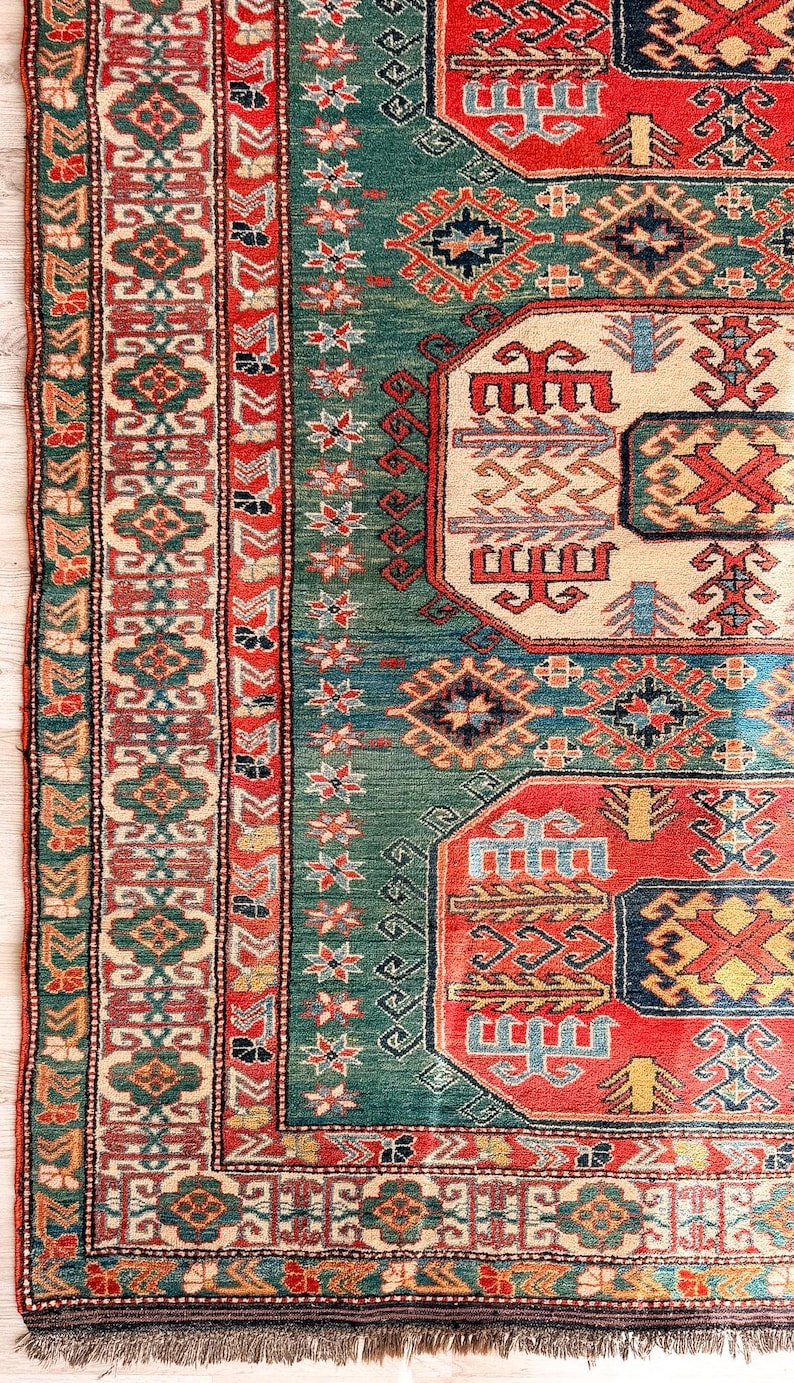 Puede incluir: Una alfombra rectangular con un campo verde intenso y medallones geom&eacute;tricos repetitivos en rojo, crema y azul. El borde muestra intrincados patrones florales y en zigzag en tonos rojos, verdes y crema, rematada con un fleco corto.