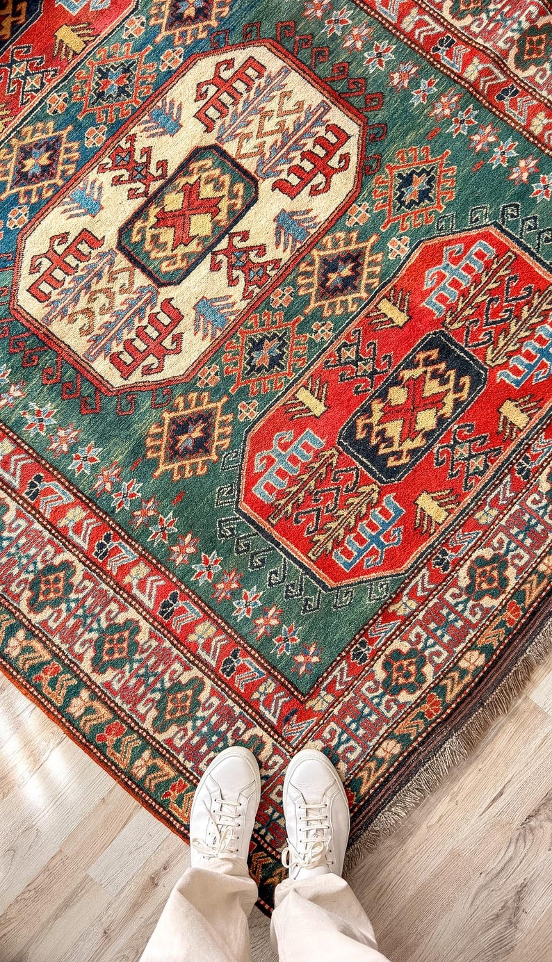Puede incluir: Una alfombra rectangular con un complejo patr&oacute;n geom&eacute;trico en tonos rojo intenso, verde bosque, azul marino y crema. El dise&ntilde;o incluye bordes intrincados y medallones centrales con motivos estilizados, rematada con un borde de flecos cortos.
