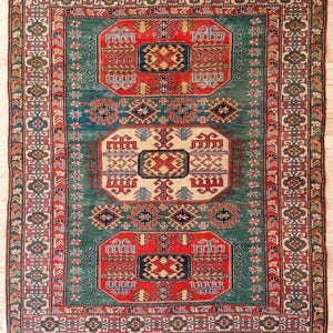Puede incluir: Una alfombra rectangular con un campo verde intenso y tres medallones centrales prominentes en rojo y crema. La alfombra presenta intrincados patrones geom&eacute;tricos, motivos florales y un borde de varias capas en rojo, verde y azul con flecos.