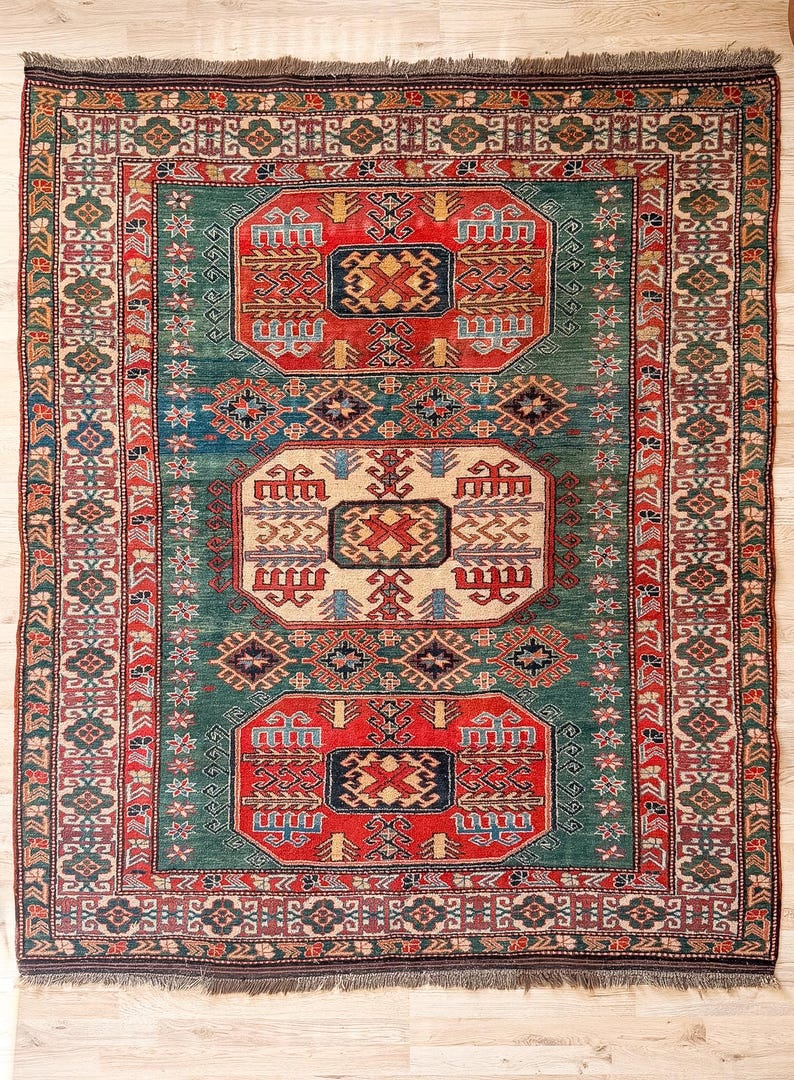 Puede incluir: Una alfombra rectangular con un campo verde intenso y tres medallones centrales prominentes en rojo y crema. La alfombra presenta intrincados patrones geom&eacute;tricos, motivos florales y un borde de varias capas en rojo, verde y azul con flecos.
