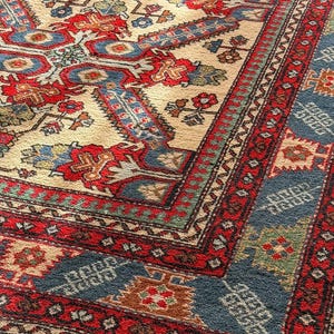Op de afbeelding: Een rechthoekig vloerkleed met een cr&egrave;mekleurige ondergrond en een centraal geometrisch medaillon in rood en blauw. De rand bevat ingewikkelde patronen in rood, blauw en groen, afgewerkt met een korte, lichtgekleurde franje aan de rand.