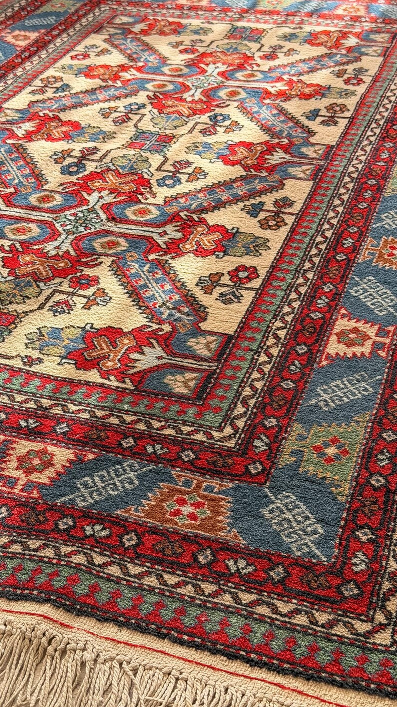 Op de afbeelding: Een rechthoekig vloerkleed met een cr&egrave;mekleurige ondergrond en een centraal geometrisch medaillon in rood en blauw. De rand bevat ingewikkelde patronen in rood, blauw en groen, afgewerkt met een korte, lichtgekleurde franje aan de rand.