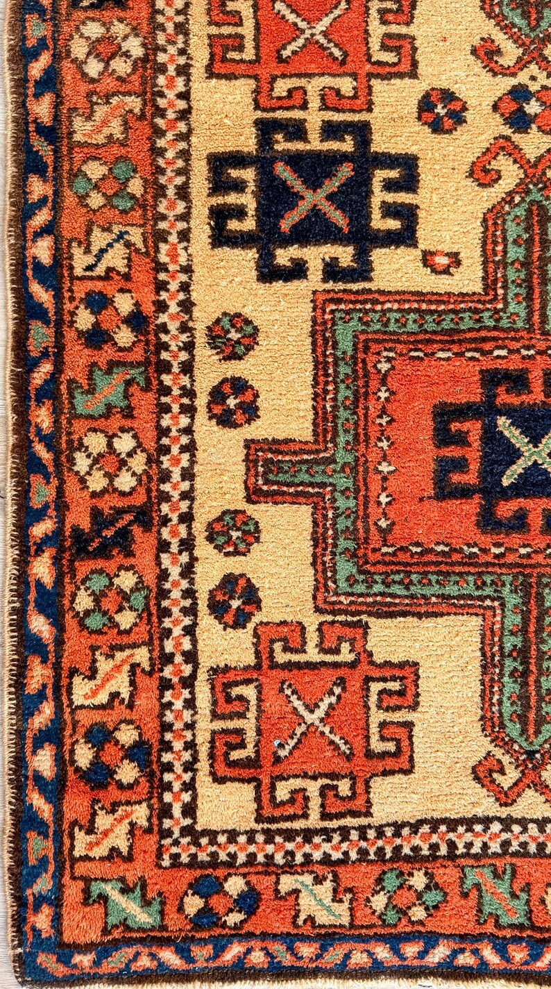 K&ouml;nnte beinhalten: Ein gewebter Teppich mit cremefarbenem Feld und geometrischen Mustern in Orange, Marineblau und Salbeigr&uuml;n. Die Bord&uuml;re zeigt ein sich wiederholendes Blumenmotiv in Orange und Blau, eingerahmt von einem dunkelblauen Rand mit gekn&uuml;pfter Struktur.