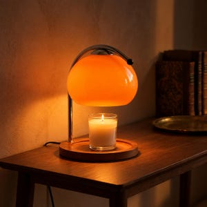 Pode incluir: Uma lâmpada aquecedora de velas com uma cúpula de vidro laranja brilhante, suportada por um braço de metal cromado. A lâmpada assenta numa base circular de madeira que contém uma vela num frasco de vidro com um pavio branco aceso.