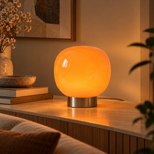 May include: A spherical table lamp featuring a smooth, translucent orange glass shade that emits a warm glow. The lamp is supported by a cylindrical, brushed silver-toned metal base, providing a stable foundation for the rounded glass structure.