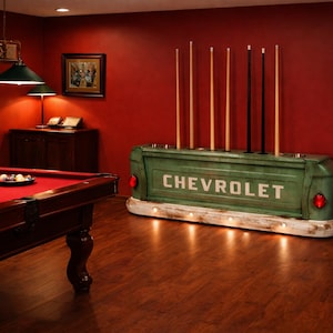Op de afbeelding: Een biljartkamer met een rode vilten pooltafel, keuen en een groene Chevrolet truckbed keuenrek. De kamer heeft een warme sfeer met donkere houten vloeren en rode muren.