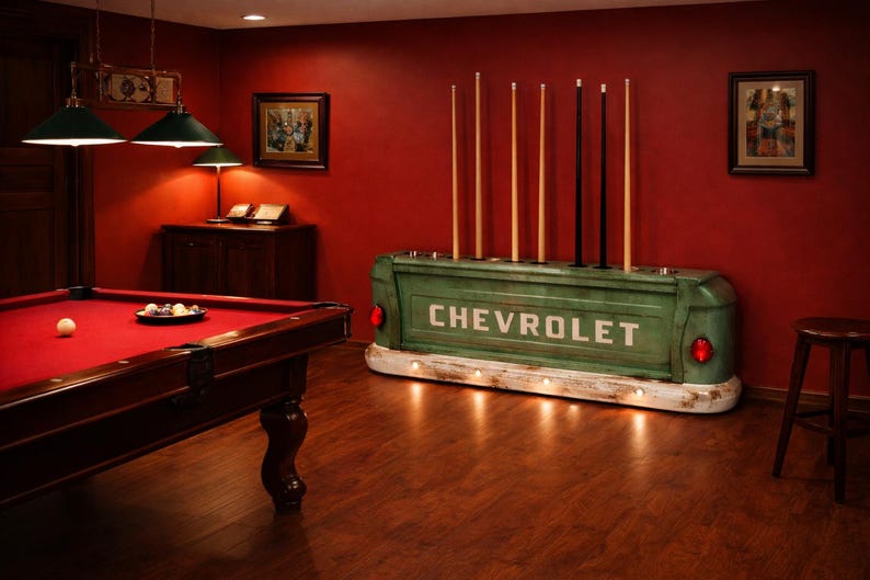 Op de afbeelding: Een biljartkamer met een rode vilten pooltafel, keuen en een groene Chevrolet truckbed keuenrek. De kamer heeft een warme sfeer met donkere houten vloeren en rode muren.